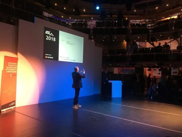 Foto: Katy Arnold: Per Axbom på scen tar ett foto av publiken, med bilden för Camp Digital 2018 bakom sig.