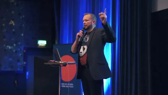 Per Axbom på en mörk scen, pekar uppåt med en mikrofon i andra handen.