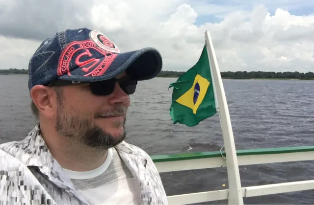 Närbild av Per Axbom i keps och solglasögon på en båt, med vatten bakom sig och land i fjärran.