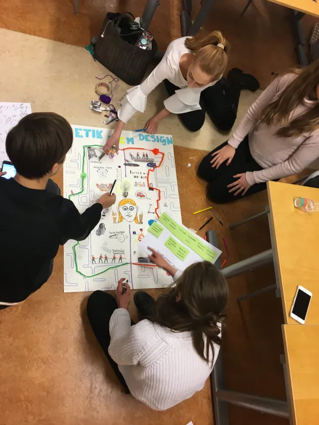 Studenter i arbete, februari 2019: Fyra studenter sitter på knä runt ett stort papper på golvet och ritar och skriver en affisch med rubriken ”Etik inom design”.