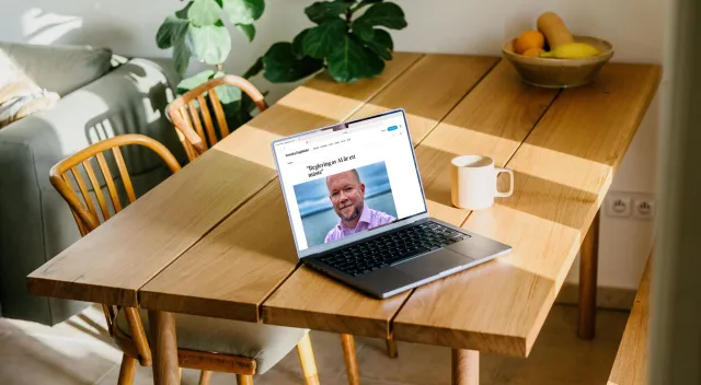 En laptop på ett solbelyst köksbord i trä, bredvid en kaffemugg och en fruktskål. På skärmen: Svenska Dagbladets debattsida med rubriken ”Reglering av AI är ett måste” och ett foto av Per.
