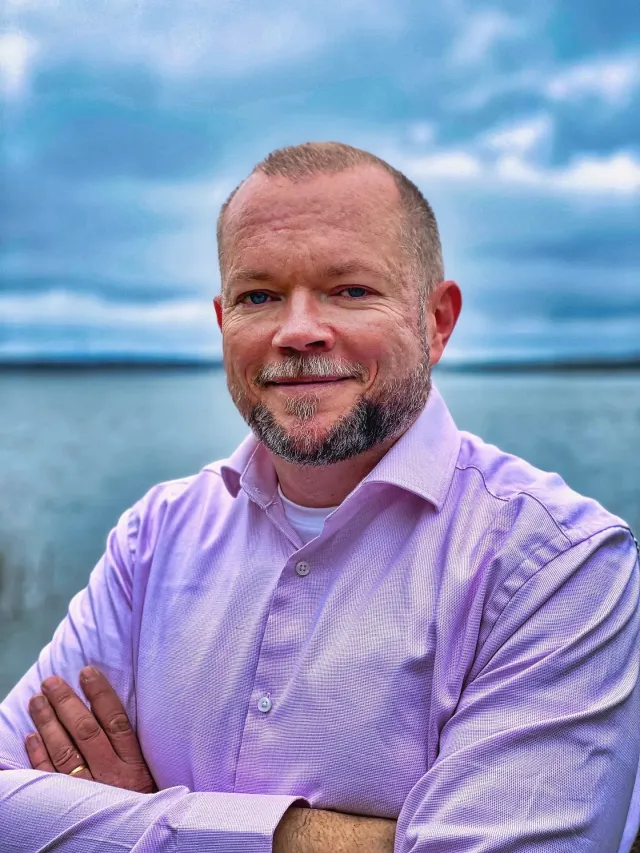 Foto: Simon Axbom: Porträttet som publicerades med artikeln: Per i lila skjorta, med armarna i kors, leende, vid en sjö under en molnig blå himmel.
