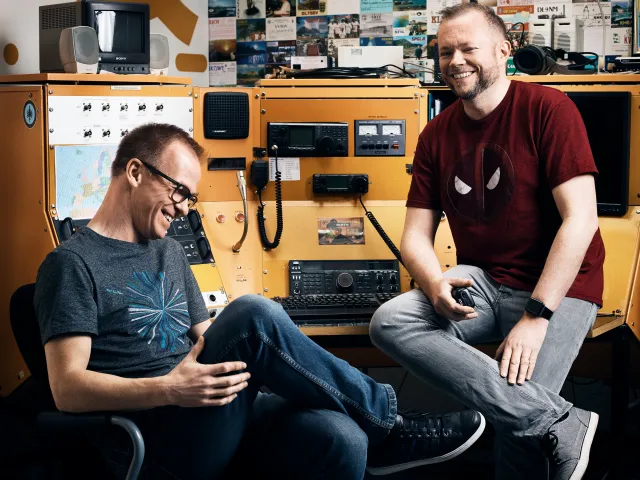 Per Axbom (till höger, i röd t-shirt) och James Royal-Lawson skrattar vid ett orange radiobord fullt av amatörradioutrustning.
