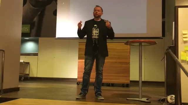 Stillbild ur inspelningen: Per på scenen i jeans, mörk kavaj och t-shirt, gestikulerar med båda händerna, med en tom projektorduk bakom sig – föredraget hölls utan bilder.